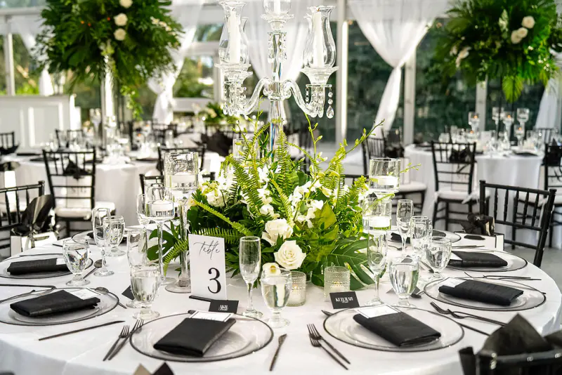 Reception table set for dinner with glassware, black napkins and a white rose and fern centerpiece under a clear tent